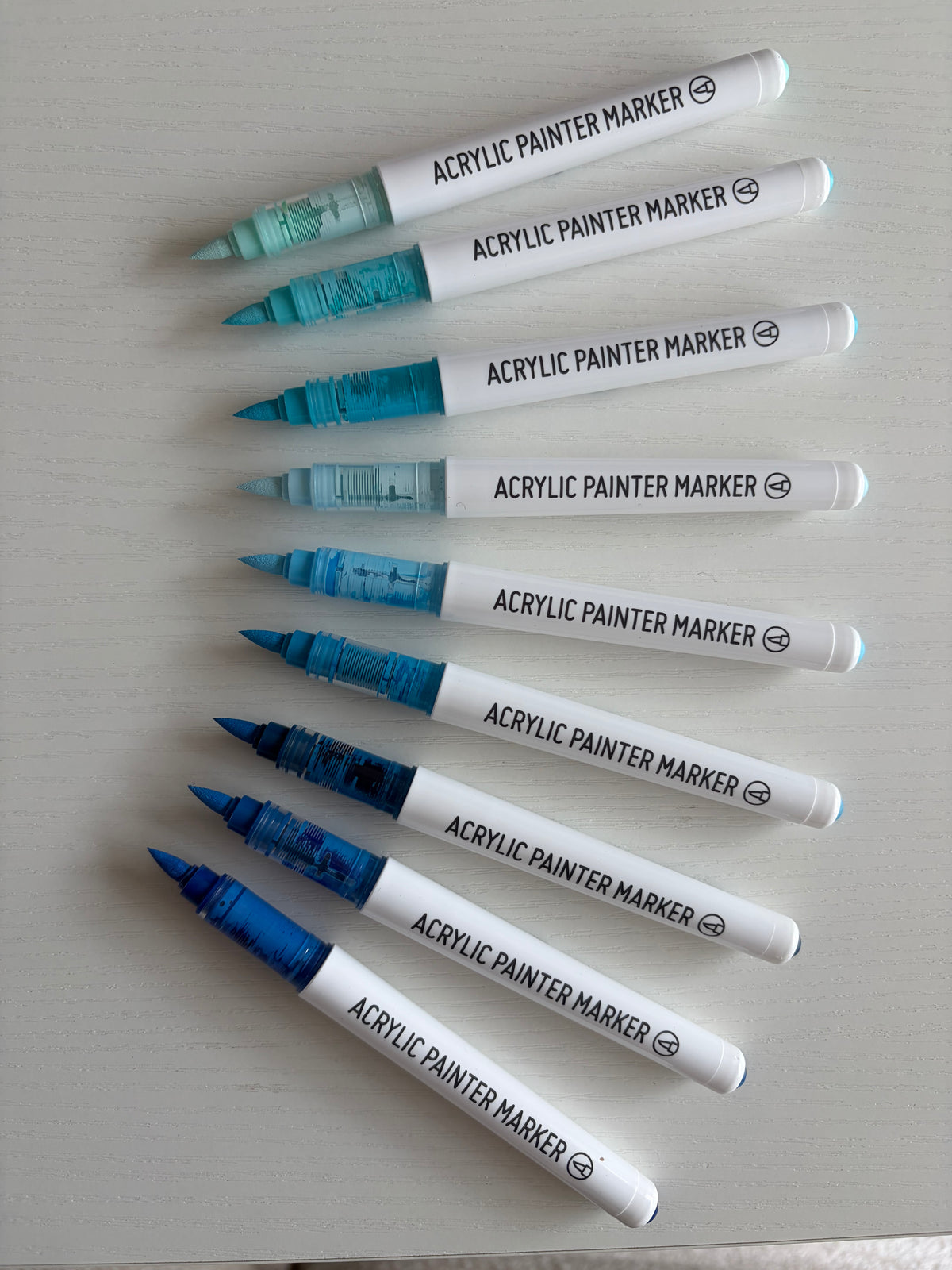 Nine acrylic paint markers with white barrels and blue/teal tips arranged diagonally on a light gray surface. The markers show different ink shades from pale blue to deep royal blue.