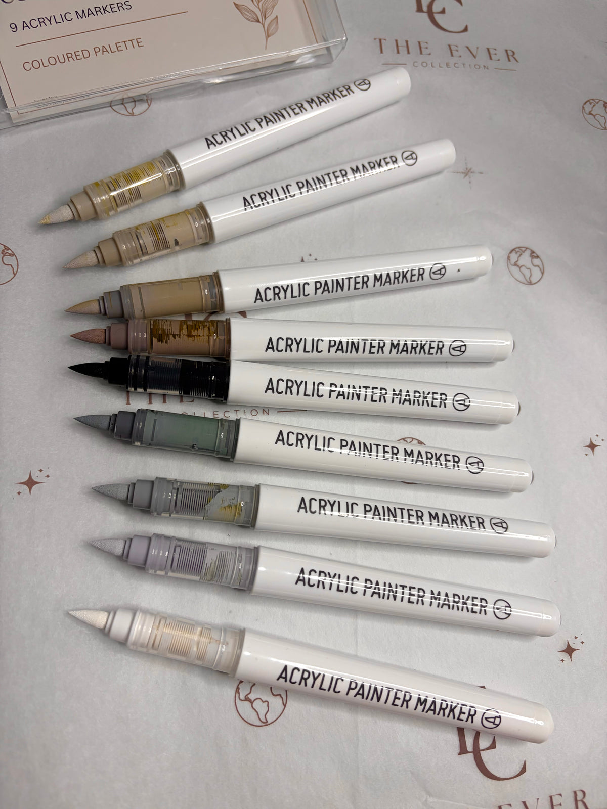 Nine acrylic paint markers from The Ever Collection arranged diagonally on a branded white background, with a small package card visible in the top left corner.