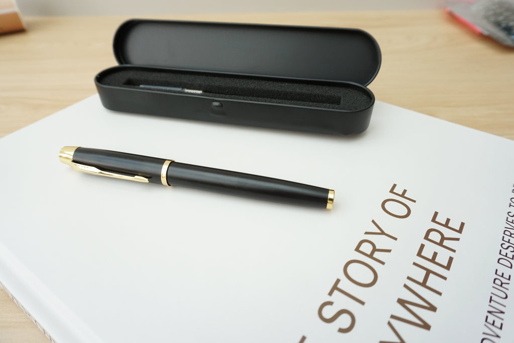 A black fountain pen with gold trim resting on a white book, with its matching black case open behind it on a light wood surface.