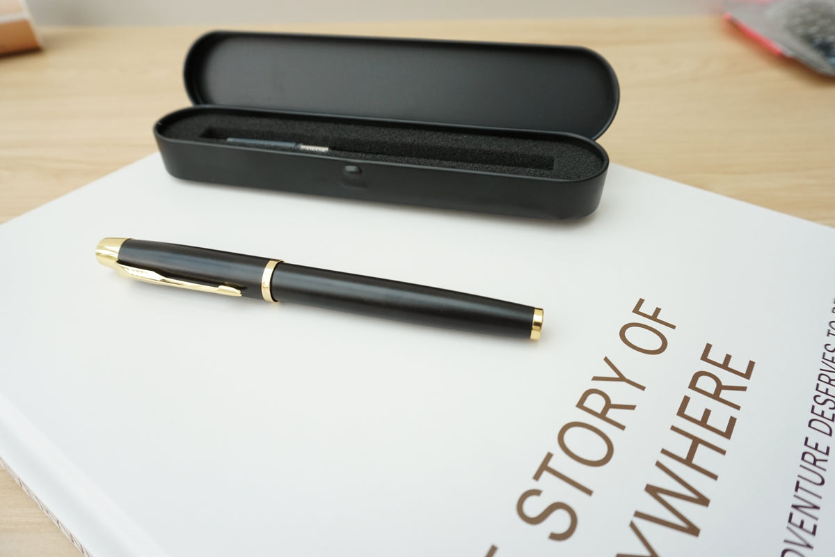 A black fountain pen with gold trim resting on a white book, with its matching black case open behind it on a light wood surface.