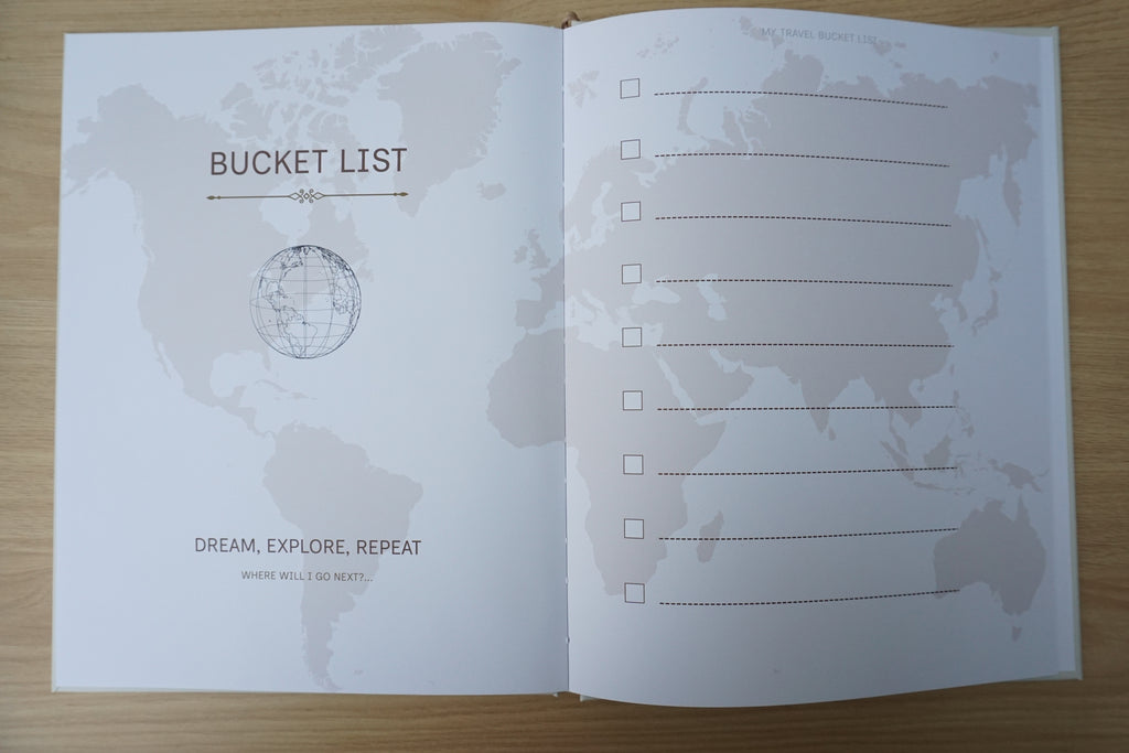 An open printed bucket list journal or booklet laid flat on a light wood surface. The left page has the title 'BUCKET LIST' above a globe illustration and the phrase 'DREAM, EXPLORE, REPEAT WHERE WILL I GO NEXT?'. Right side shows a check list to be filled in 