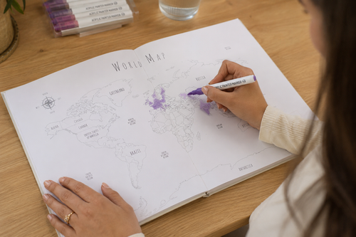 A person coloring a world map book on a wooden desk with a purple marker.