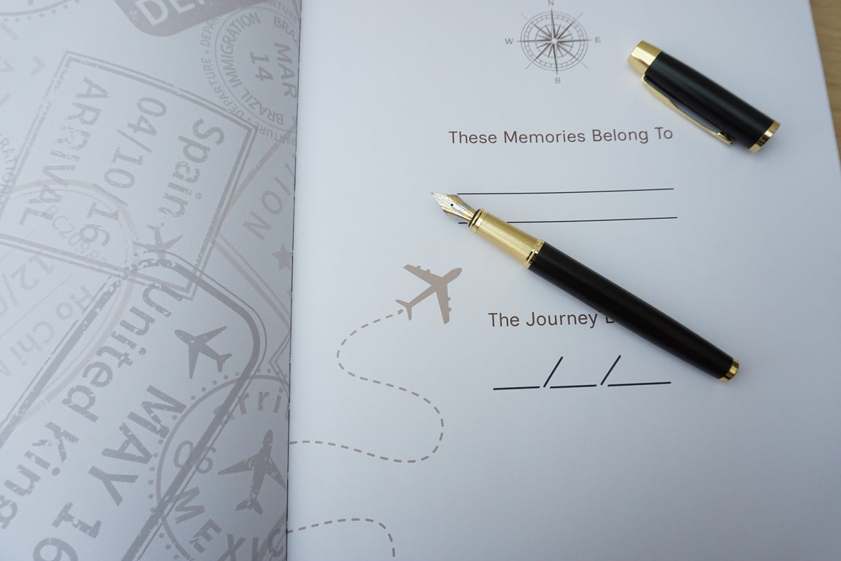 A black fountain pen with gold trim and a matching pen cap rests on a white memorial journal page with travel-themed illustrations, next to a passport-style stamp booklet.