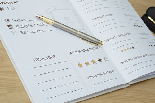 An open white journal or planner on a light wood desk, featuring printed prompts and sections such as Adventure, Highlight, and Adventure Rating. A gold and silver pen rests across the pages, with a black case partially visible on the right.